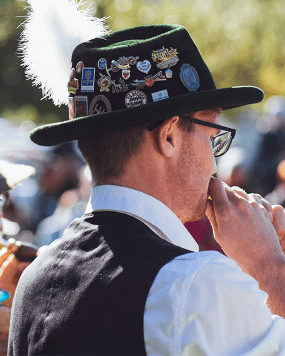 Mitglied eines bayerischen Trachtenvereins bei einem traditionellen Volksfest. Er trägt einen Hut mit Abzeichen und Feder sowie Weste und Hemd, während er ein Blasinstrument spielt.