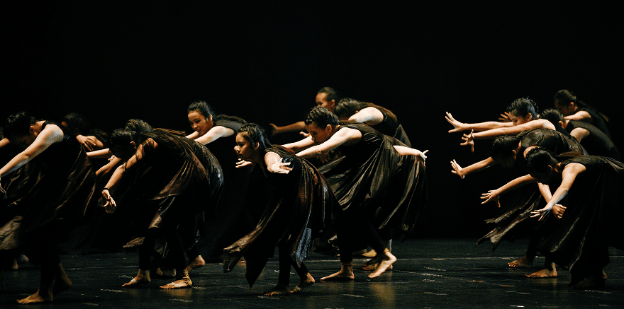 Ensemble von Tänzer:innen in dunklen Kostümen während einer zeitgenössischen Tanzperformance auf einer Bühne in Bayern. Die Gruppe bewegt sich synchron und ausdrucksstark im Scheinwerferlicht eines Theatersaals.